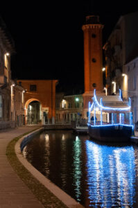 Night view of the canal with the Torre Civica and a restaurant boat - Comacchio, Italy - rossiwrites.com