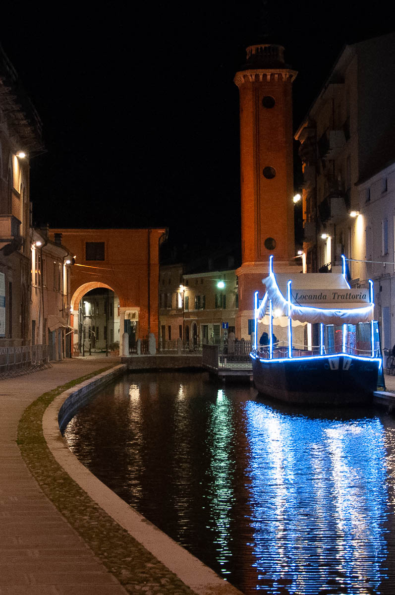 Night view of the canal with the Torre Civica and a restaurant boat - Comacchio, Italy - rossiwrites.com